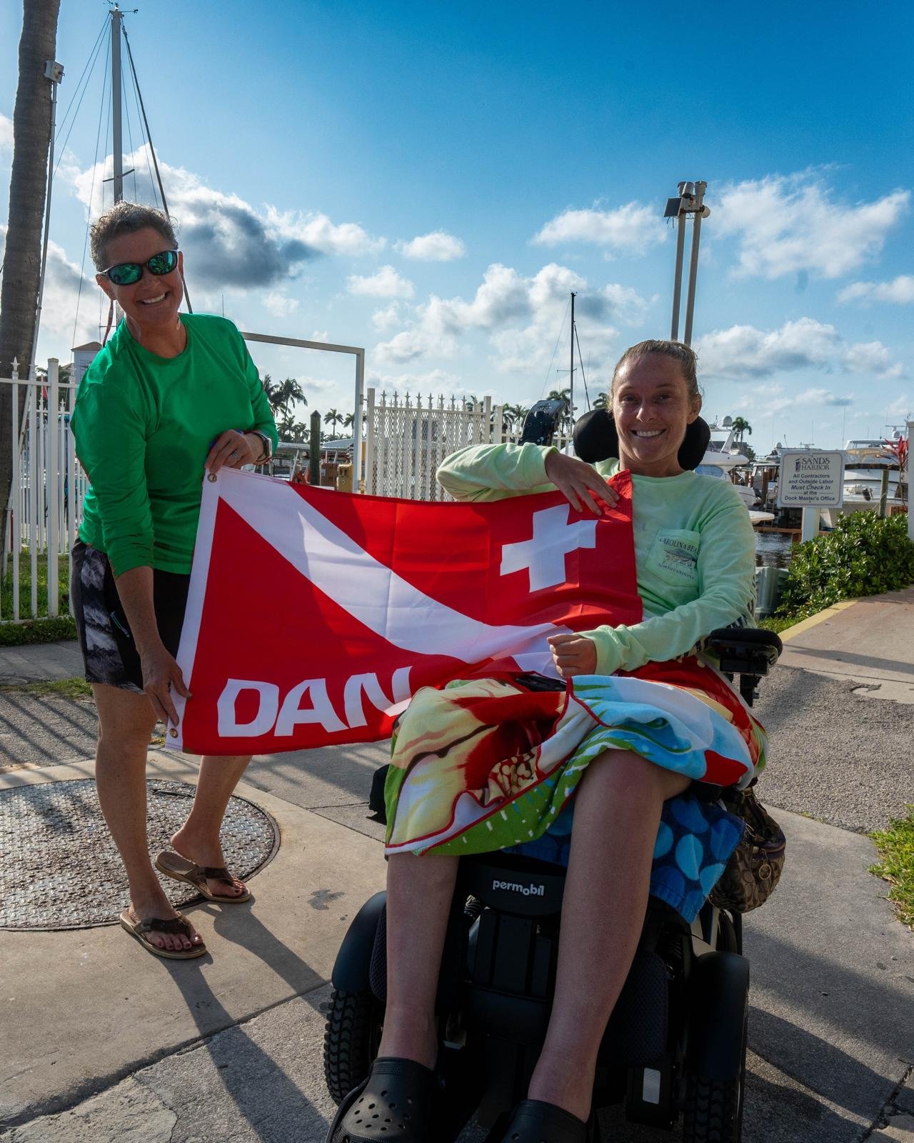 Two women smile while holding a red DAN dive flag, one seated in a power wheelchair with a beach towel draped across her lap at a sunny marina.